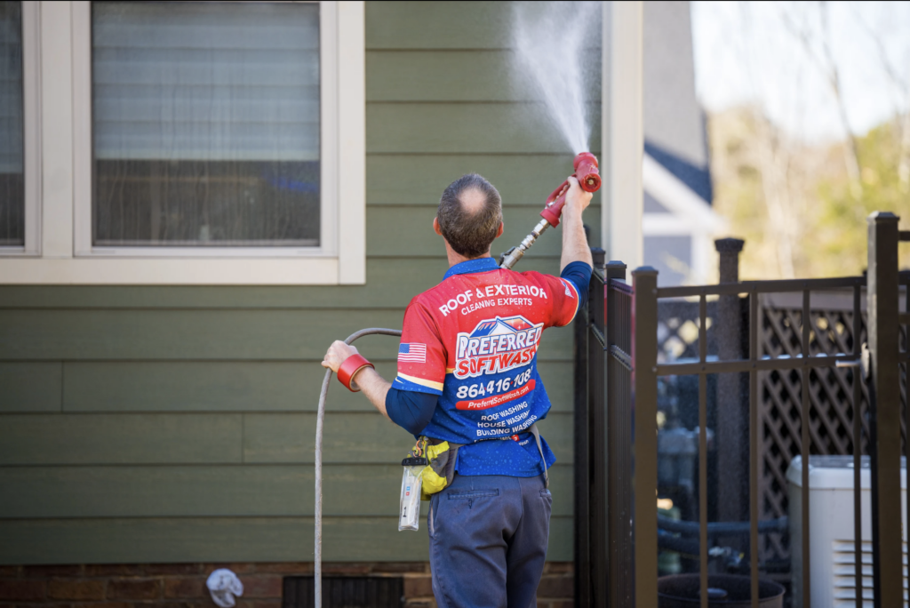 A person in a red and blue uniform is pressure washing the exterior wall of a house. The uniform has various texts and logos on the back. The person is holding a hose with water spraying onto the green wooden siding of the house. There is a black fence nearby.